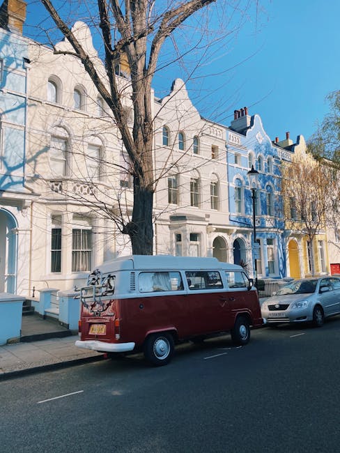 A classic red and white vintage van parked on a residential street in Notting Hill, with a ladder and other equipment secured on the roof. Behind the van, a row of colorful Victorian-style terraced houses with ornate facades and arched windows can be seen, some with blue and yellow paintwork. The street has parked cars alongside the van, and a large leafless tree extends branches overhead. The scene captures a typical urban environment in London, with clear blue skies and bright daylight, during a home relocation or furniture transport process involving removals services, such as those provided by Removals Notting Hill.