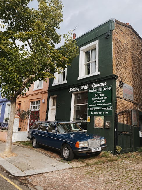 The image shows a two-storey green building on a narrow street in Notting Hill, with a sign indicating it is the Notting Hill Garage offering car storage and repair services. A vintage dark blue station wagon with a roof rack is parked directly in front of the garage on the cobblestone pavement, partially blocking the building's entrance. To the left, a tree with green foliage provides some shade, and a neighboring peach-coloured building is visible further along the street. The scene is captured during daylight with overcast skies, and the adjacent street features adjacent residential buildings with mixed architectural styles. This setting illustrates typical London street scenery, relevant to house removals and moving services, where parking restrictions and vehicle placement are key considerations during a home relocation handled by companies like Removals Notting Hill.