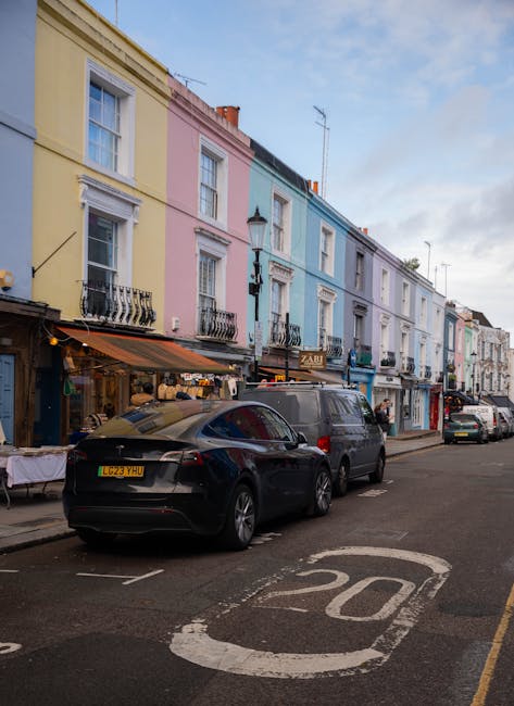 A classic red and white vintage van parked on a residential street in Notting Hill, with a ladder and other equipment secured on the roof. Behind the van, a row of colorful Victorian-style terraced houses with ornate facades and arched windows can be seen, some with blue and yellow paintwork. The street has parked cars alongside the van, and a large leafless tree extends branches overhead. The scene captures a typical urban environment in London, with clear blue skies and bright daylight, during a home relocation or furniture transport process involving removals services, such as those provided by Removals Notting Hill.