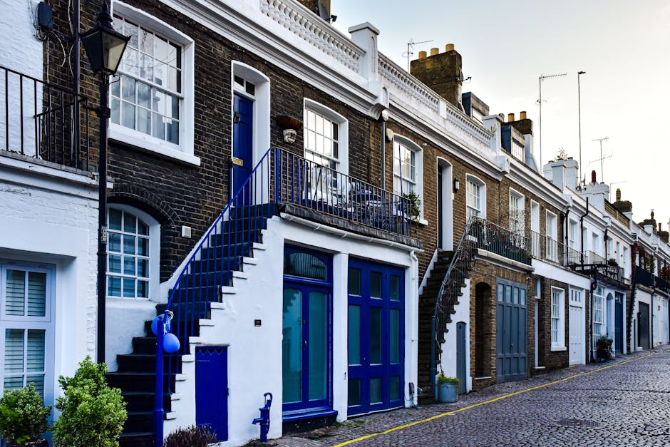 A row of terraced houses with dark brick facades and white window frames on a cobbled street in Ladbroke Grove, during daytime with clear weather. The houses feature external staircases with black met