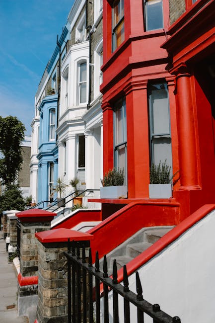 A row of residential houses featuring a mix of white and red facades with large windows and decorative architectural elements visible on a residential street. The red building in the foreground has a concrete staircase with a metal railing leading up to a front door, flanked by two potted plants in rectangular white containers. The pavement and steps appear clean, and there are clear skies overhead. Visible in the image is the exterior of the building, with some shadowing suggesting natural daylight. The scene captures the typical urban environment for house removals and furniture transport, as [COMPANY_NAME] facilitates home relocation services in this area, which often involves careful packing and loading processes to move bulky furniture efficiently without causing damage or fines when clearing properties.