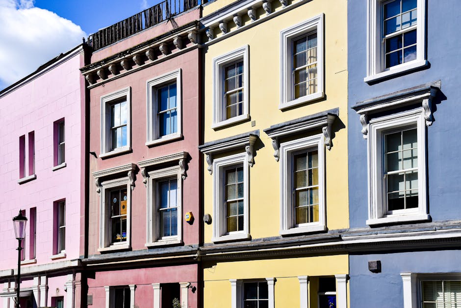 A row of three residential buildings with pastel-colored facades in pink, yellow, and blue, each featuring large white-framed sash windows and decorative molding. The buildings are multi-storey, with the pink building on the left, the yellow in the middle, and the blue on the right. In the foreground, a street lamp is visible on the pavement. The sky above is partly cloudy with patches of blue. This exterior scene relates to house removals and moving services focused on the logistical process of clearing bulky furniture in Notting Hill without the need for council fines, as provided by Removals Notting Hill.