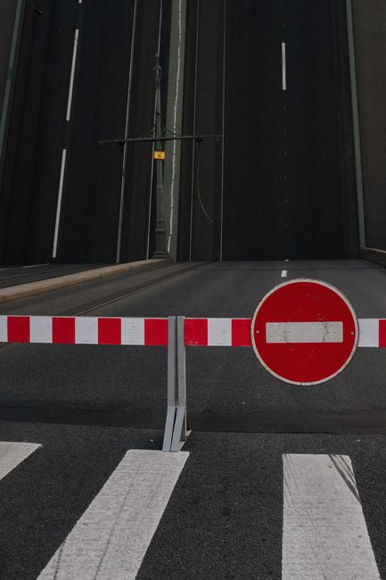 A road closure barrier with a red and white striped horizontal bar and a 'no entry' circular sign positioned across a street in front of a modern building with dark exterior panels. The barrier is placed on the asphalt, blocking vehicle access to the entrance, which features a large, dark gate or door. The surrounding area includes a pedestrian crossing with white striped markings on the pavement in the foreground. The scene appears to be part of traffic management measures, possibly related to house removals or relocation services in Notting Hill, as indicated by the context of the page about low-loader and road closure regulations. The lighting suggests daytime, with no visible vehicles or people present.