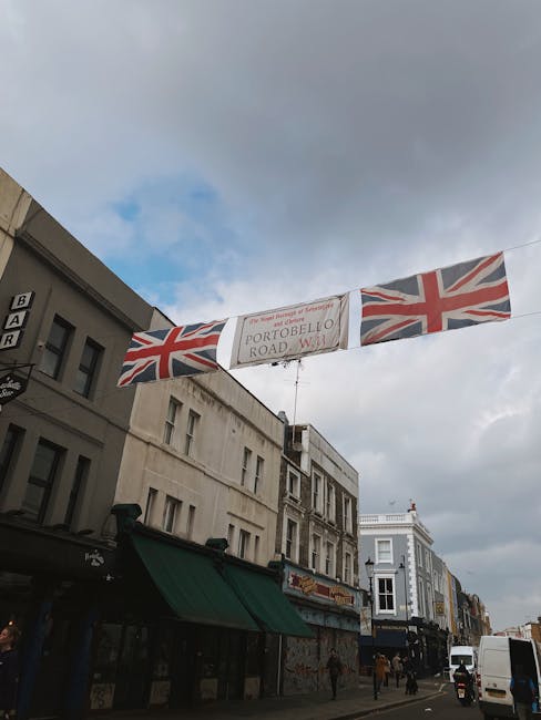 Photograph of a city street in Notting Hill, showing a banner suspended across the road with a British Union Jack flag on each side and a central sign reading 'Portobello Road W11'. The street features various buildings, including shops and residences, with some signage and awnings visible. In the foreground, a white van is parked near the curb, and a few pedestrians are walking along the pavement. The sky is partly cloudy, providing diffused natural lighting. This scene reflects typical urban activity associated with house removals and moving logistics in the area, where careful planning behind road closures and regulations is essential for home relocation and furniture transport, as highlighted by the contents of the page on low-loader and road closure regulations for Notting Hill at removalsnottinghill.com.