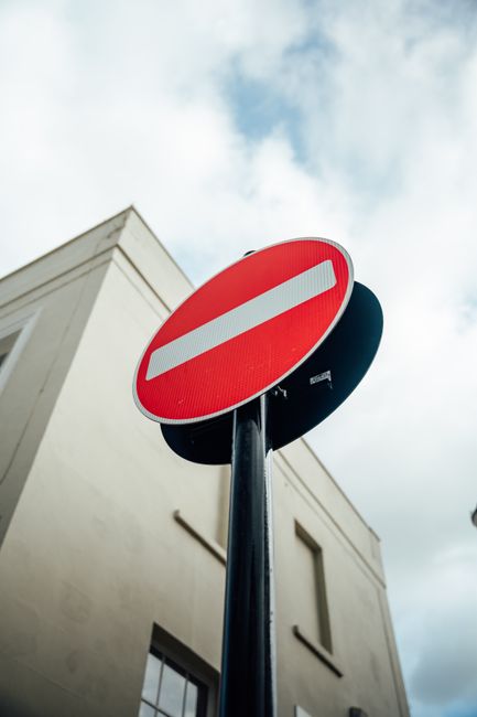 A road closure barrier with a red and white striped horizontal bar and a 'no entry' circular sign positioned across a street in front of a modern building with dark exterior panels. The barrier is placed on the asphalt, blocking vehicle access to the entrance, which features a large, dark gate or door. The surrounding area includes a pedestrian crossing with white striped markings on the pavement in the foreground. The scene appears to be part of traffic management measures, possibly related to house removals or relocation services in Notting Hill, as indicated by the context of the page about low-loader and road closure regulations. The lighting suggests daytime, with no visible vehicles or people present.