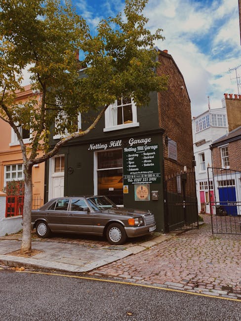 The exterior of a multi-storey building housing Notting Hill Garage, a home relocation and furniture transport service, with a dark green facade and signage listing services such as removals, packing, storage, and furniture lifts. A classic grey sedan is parked on a cobblestone street directly in front of the garage entrance. The street is lined with a mature tree, with green leaves partially shading the vehicle, and a black metal fence borders the property. In the background, neighboring buildings with white and red brick facades are visible under a partly cloudy sky, with some residential balconies seen on the upper floors. The scene depicts a typical urban setting for furniture loading and unloading activities associated with professional removals and house moving in the Notting Hill area, aligning with the page titled 'Notting Hill Gate narrow-stair removals: safe packing & lifting' at removalsnottinghill.com. The process hints at a loading process for estate clearance or home relocation, emphasizing careful handling and organized transport.