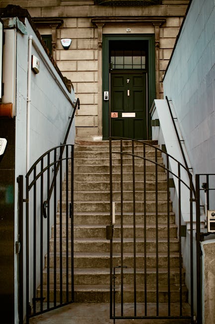 The image shows a set of exterior concrete stairs leading up to a dark green wooden front door of a residential building, which features a rectangular window with a grid pattern and a mailbox slot below. The stairs are bordered by black wrought iron railings on both sides, with a small gate at the bottom, also made of black iron. To the left of the stairs, a tall, light grey metal fence extends upward, with a small security camera mounted near the top. To the right, there is a white, plastic-covered panel or sign attached to the building wall, which appears to be part of the building's utility infrastructure. The scene is well-lit with natural daylight, capturing the typical setting for a home relocation process, such as loading or unloading furniture and moving boxes, in front of a classic London-style townhouse. This image exemplifies the logistical considerations seen during house removals and packing for a safe and efficient move, which Removals Notting Hill professionally provides in the Notting Hill area.