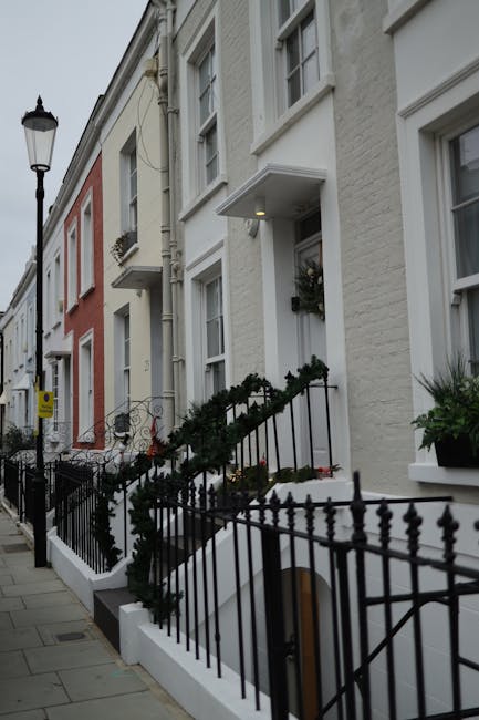The exterior of a multi-storey building housing Notting Hill Garage, a home relocation and furniture transport service, with a dark green facade and signage listing services such as removals, packing, storage, and furniture lifts. A classic grey sedan is parked on a cobblestone street directly in front of the garage entrance. The street is lined with a mature tree, with green leaves partially shading the vehicle, and a black metal fence borders the property. In the background, neighboring buildings with white and red brick facades are visible under a partly cloudy sky, with some residential balconies seen on the upper floors. The scene depicts a typical urban setting for furniture loading and unloading activities associated with professional removals and house moving in the Notting Hill area, aligning with the page titled 'Notting Hill Gate narrow-stair removals: safe packing & lifting' at removalsnottinghill.com. The process hints at a loading process for estate clearance or home relocation, emphasizing careful handling and organized transport.