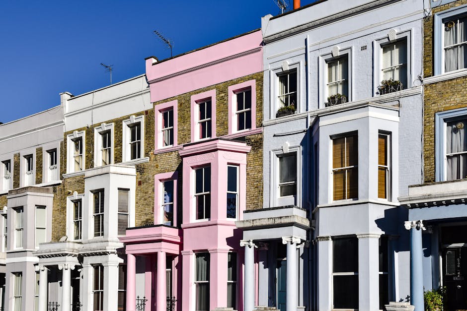 Exterior view of a row of Victorian-style terraced houses in Notting Hill, London, showcasing a mix of white, pink, and light blue facades. The buildings feature large sash windows, some with flower boxes, and decorative architectural details such as bay windows and cornices. In the foreground, a home relocation process is underway with furniture and cardboard boxes visibly being loaded onto a black moving van parked on the pavement, partially visible behind the houses. The loading area is illuminated by bright daylight, highlighting the materials used in packing and transport, such as plastic wrapping and moving blankets. A trolley and straps are used to assist with the heavy lifting of furniture, and the process is managed carefully by Removals Notting Hill as part of professional furniture transport and packing services to ensure a smooth house move and clear understanding of removal pricing, as explained on their website. The scene captures both the visually appealing architecture and the logistical aspects of home relocation, including careful handling of belongings during the loading process for residential moves in the area.