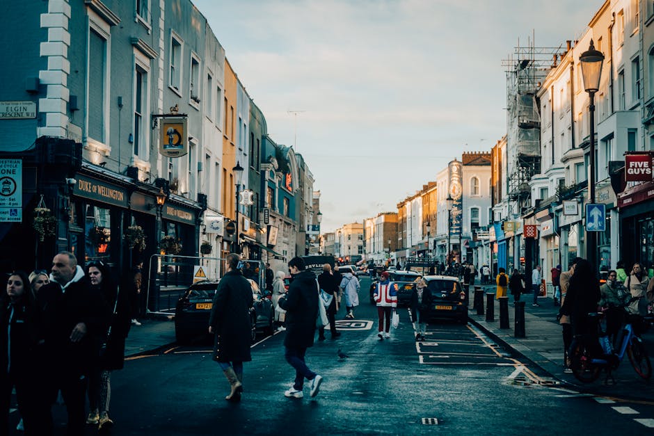 A busy street scene in Notting Hill during the late afternoon, featuring a row of commercial buildings with shops and cafes on both sides. The street is filled with pedestrians walking along the pavement, some carrying shopping bags, and a few standing near the shop entrances. Several parked cars and taxis are aligned along the curb, with a few vehicles moving through the street. A man in a red and white high-visibility vest stands near the center of the street, possibly involved in loading or directing traffic. Street lamps and signs are visible, with scaffolding on some of the buildings indicating ongoing maintenance or renovations. The natural daylight creates a warm glow on the buildings, and the overall scene captures the lively, urban atmosphere typical of a home relocation or moving logistical environment, aligning with the services offered by Removals Notting Hill.