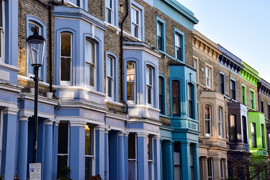 Photograph of a row of Victorian-style terraced houses in Notting Hill with diverse coloured facades, including light blue, beige, bright green, and navy. Each building features large sash windows, some with decorative molding and window arches. The ground floor windows are partially visible, and a black street lamp stands in front of the houses on a pavement lined with a small parking sign. The scene is captured in natural daylight, with clear blue skies overhead. This visual illustrates the typical exterior of residential properties in the area, relevant to home relocation and furniture transport services provided by Removals Notting Hill, who assist with packing, moving, and loading objects during house removals in this neighbourhood.