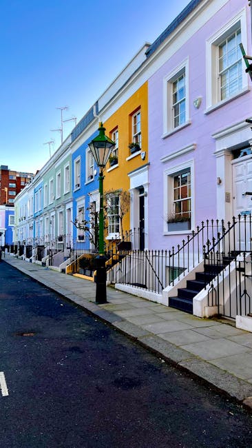 A busy street scene in Notting Hill during the late afternoon, featuring a row of commercial buildings with shops and cafes on both sides. The street is filled with pedestrians walking along the pavement, some carrying shopping bags, and a few standing near the shop entrances. Several parked cars and taxis are aligned along the curb, with a few vehicles moving through the street. A man in a red and white high-visibility vest stands near the center of the street, possibly involved in loading or directing traffic. Street lamps and signs are visible, with scaffolding on some of the buildings indicating ongoing maintenance or renovations. The natural daylight creates a warm glow on the buildings, and the overall scene captures the lively, urban atmosphere typical of a home relocation or moving logistical environment, aligning with the services offered by Removals Notting Hill.