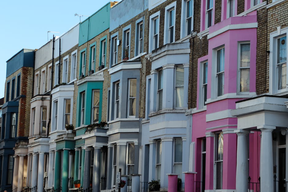 A row of Victorian terraced houses with multiple floors, each featuring large sash windows and decorative bay windows. The buildings are painted in various pastel colours, including shades of blue, pink, and white, with some brickwork visible on the upper sections. The image is taken during daylight with clear sky, showing the facades facing the street. The lower parts of the buildings have small front gardens or operate as entryways, some with potted plants or decorative elements. The scene depicts a typical residential street suitable for home relocations and furniture transport services provided by Removals Notting Hill, with a focus on the architecture and urban environment involved in house removals and packing or loading processes.