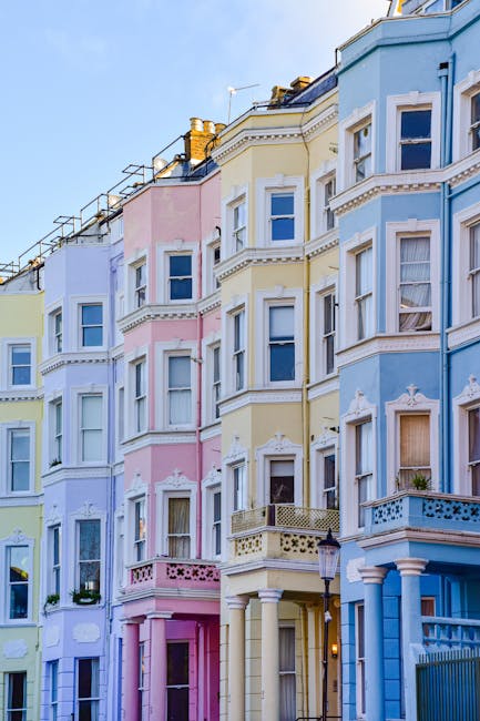 Photograph of a row of colorful Victorian terraced houses in Notting Hill, London, with each building painted in pastel shades including blue, pink, yellow, and green. The buildings feature large bay windows, some with decorative mouldings, and small front balconies. The image captures the upper floors and rooftops, with visible chimneys and roofing materials. The scene is illuminated by natural daylight, highlighting the architectural details and vibrant colors. This exterior view aligns with house removals and moving services, illustrating a typical residential street where professional relocations by Removals Notting Hill could take place during home extensions or moving processes, with a focus on the urban environment and structural facades.