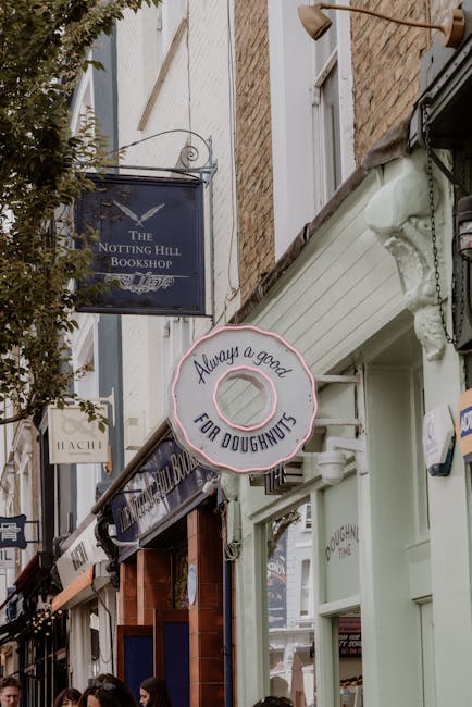 A narrow street with multiple shopfronts and signage, including a dark blue sign for The Notting Hill Bookshop and a round neon sign shaped like a doughnut with the text 'Always a good for doughnuts.' The street features brick and painted facades, with a white building having large windows and a white awning to the right. Above, there is a beige canopy extending over the sidewalk, partially covering the lower part of the storefronts. At street level, decorative elements such as hanging signs and small awnings are visible, with some pedestrians walking on the pavement. Removals Notting Hill's professional team is engaged in a home relocation or furniture transport process, visible through the presence of move-related equipment such as dollies, packing materials, or boxes being carried. The environment is well-lit with natural daylight, enhancing the visibility of the shop signage and street scene, as part of their commercial removals service in Notting Hill.
