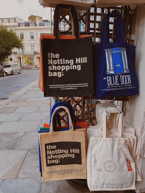 A display stand outdoors on a sidewalk, holding multiple reusable shopping bags made of fabric, some with printed logos and text. The bags include black, blue, and beige colors, featuring branding for the Notting Hill shopping bag and Blue Door Notting Hill Laundry, with handles upright and positioned to show the front designs. The stand is made of a metal grid with wooden panels, and the sidewalk is paved with square stone tiles. In the background, there are residential buildings with balconies, windows, and parked cars, indicating an urban area. The lighting appears natural, suggesting daytime. This scene reflects packing and shopping aspects related to house removals and moving services, with [COMPANY_NAME] occasionally involved in contextually supporting relocation logistics.